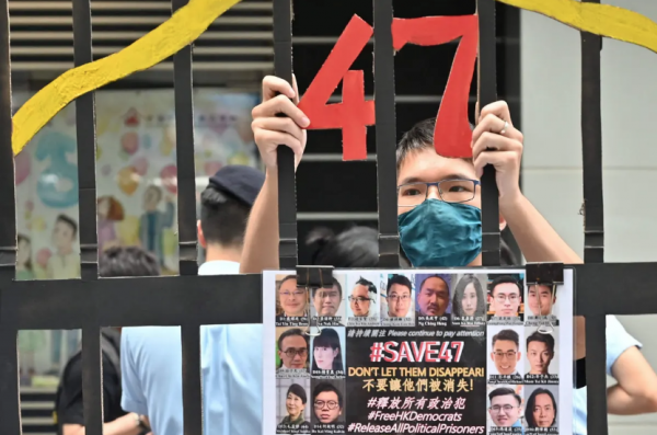 Un manifestant se tient derrière de faux barreaux de prison avec les photos des 47 figures du camp pro-democraie en procès pour "tentative de renverser le gouvernement", à Hong Kong en mai 2021. (Source : Guardian)